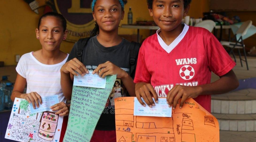 Three children in Belize show their certificates for participating in the Projects Abroad summer camp and illustrations of their work. A Projects Abroad volunteer working with children in Belize helps children celebrate with certificates they recieved for their hard work at a summer camp.